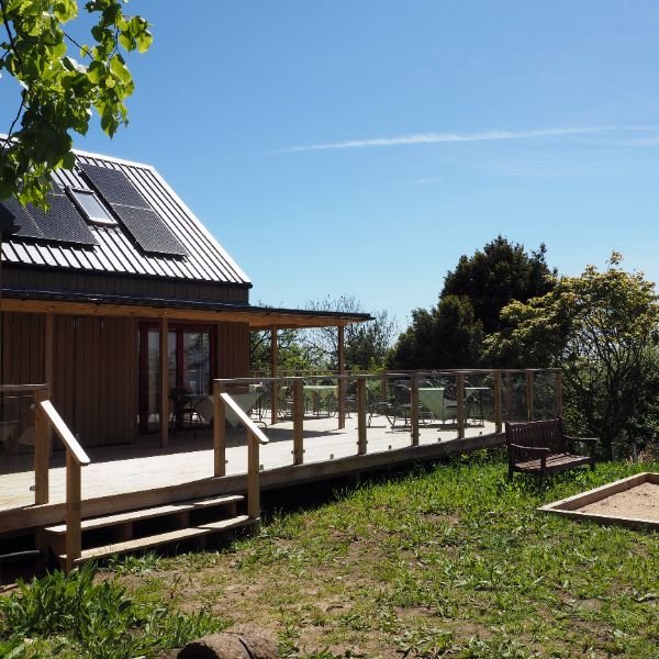 A multi-purpose studio at Othona with solar panels on a metal roof, wooden exterior, and a deck surrounded by greenery under a clear blue sky.
