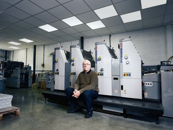 A person seated on a platform in front of a row of Heidelberg GTO 52 industrial printing machines inside a spacious, well-lit printing facility.