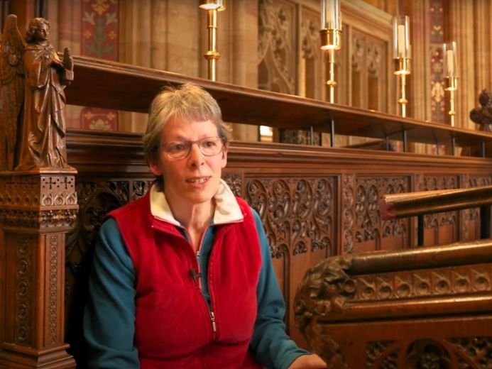 A person seated inside Sherborne Abbey, surrounded by ornate wooden carvings, statues, and tall candle holders, highlighting the historic and religious architecture.