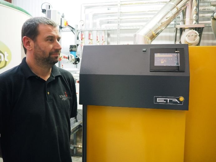 Person standing beside an ETA-branded industrial biomass machine with visible control panel and surrounding pipes.