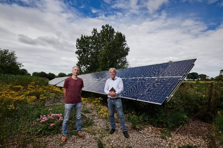 Two people standing in front of a large solar panel array at Springhead Trust, surrounded by greenery under a partly cloudy sky.