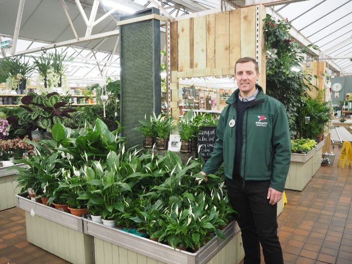 Person in a green jacket standing beside peace lilies at Stewarts Garden Centre, with plants and gardening supplies in the background.