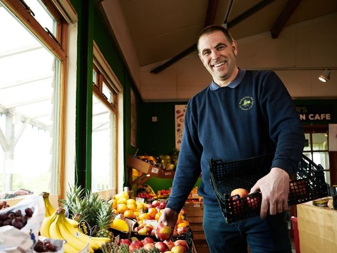 A person holding a black basket with apples stands inside a fruit and vegetable store, surrounded by displays of bananas, pineapples, grapes, and other produce, with natural light streaming through large windows.