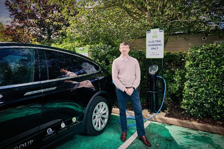 Person standing beside a black electric vehicle at a charging station in Waterside Holiday Park, with greenery in the background.