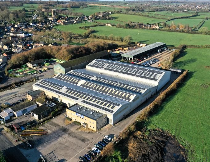 Aerial view of William Hughes industrial site with white-roofed buildings, some fitted with solar panels, surrounded by fields and a nearby village.