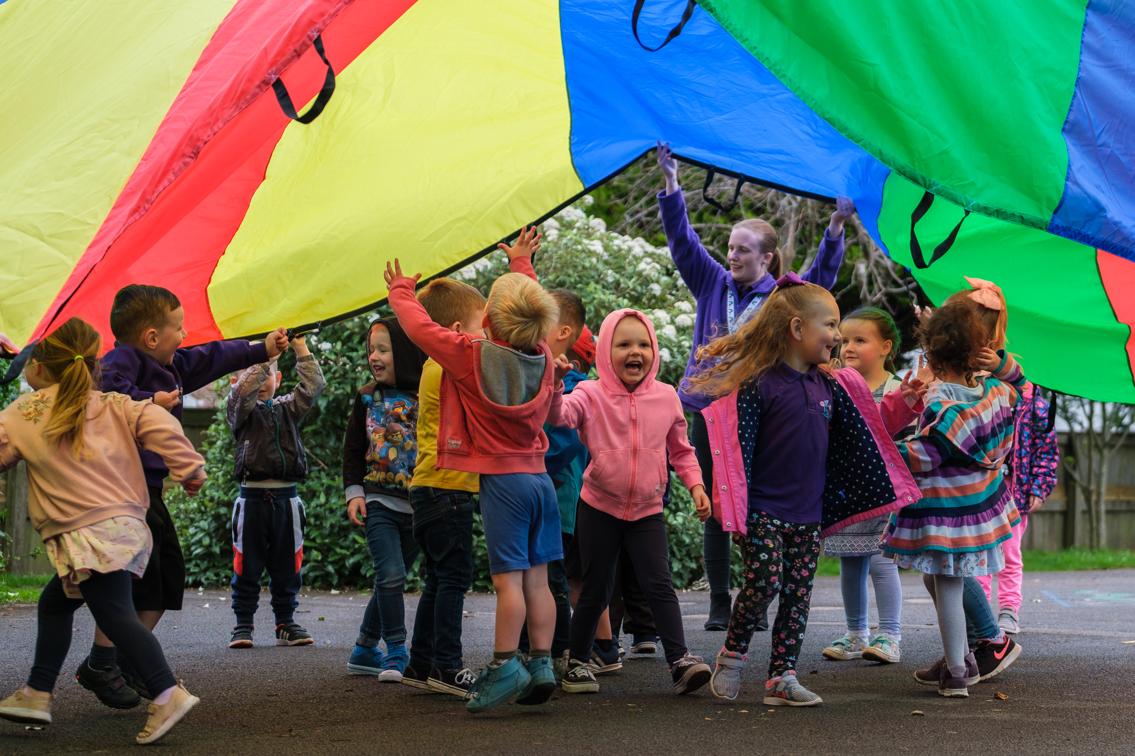 A group of young pre-school children are under a large rainbow coloured fabric circle being used for a game in a playground. There is an adult supervising, they are smiling and active.