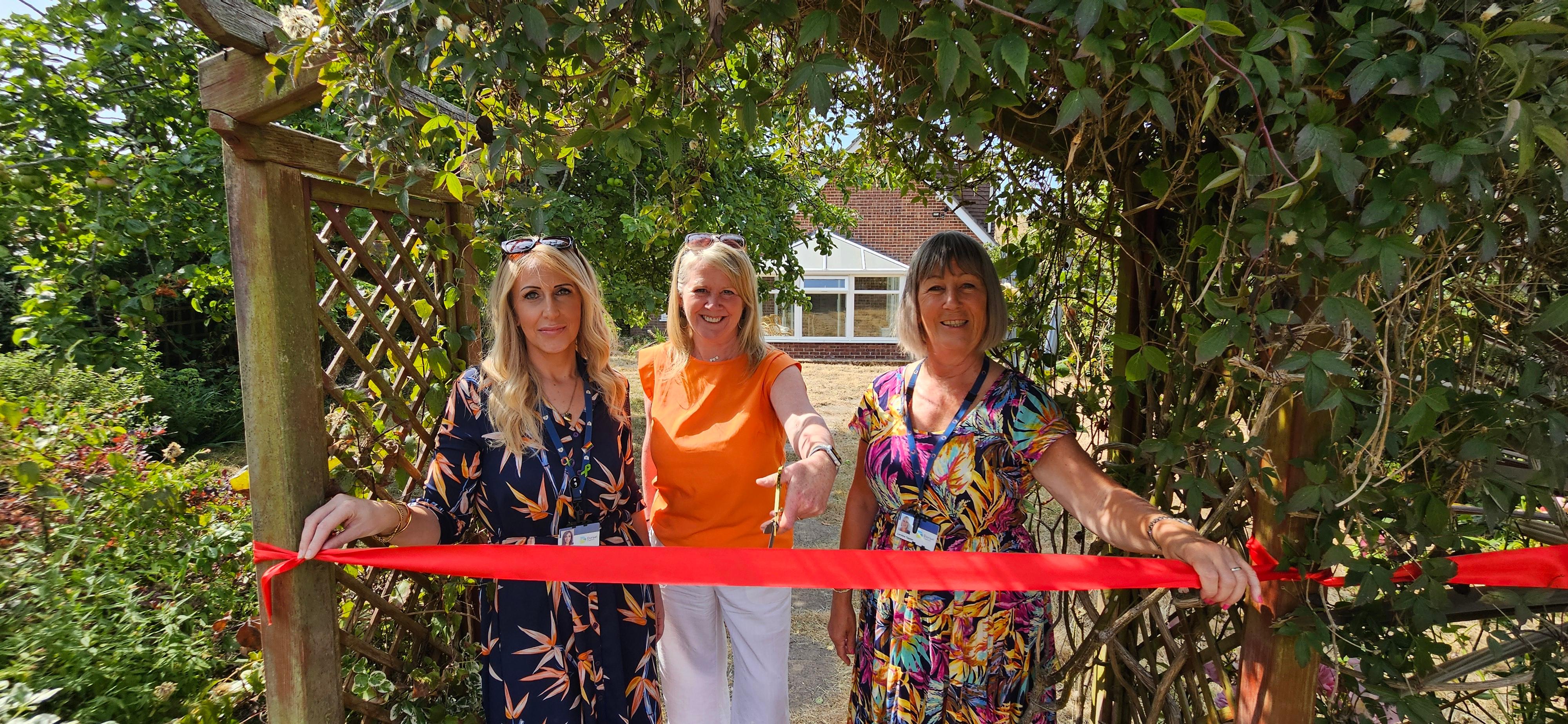 Three women stand behind a taut red ribbon which is attached to a wooden archway in a back garden. The middle woman has a pair of ceremonial scissors.