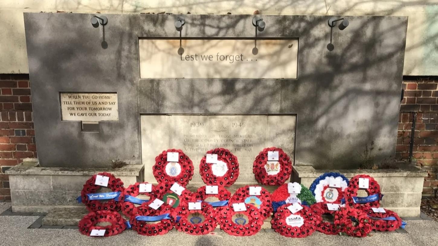 War memorial with the inscription “Lest we forget” and a commemorative plaque, surrounded by numerous red remembrance wreaths laid at its base. The memorial stands against a stone wall with tree shadows cast across the surface, and wreaths.