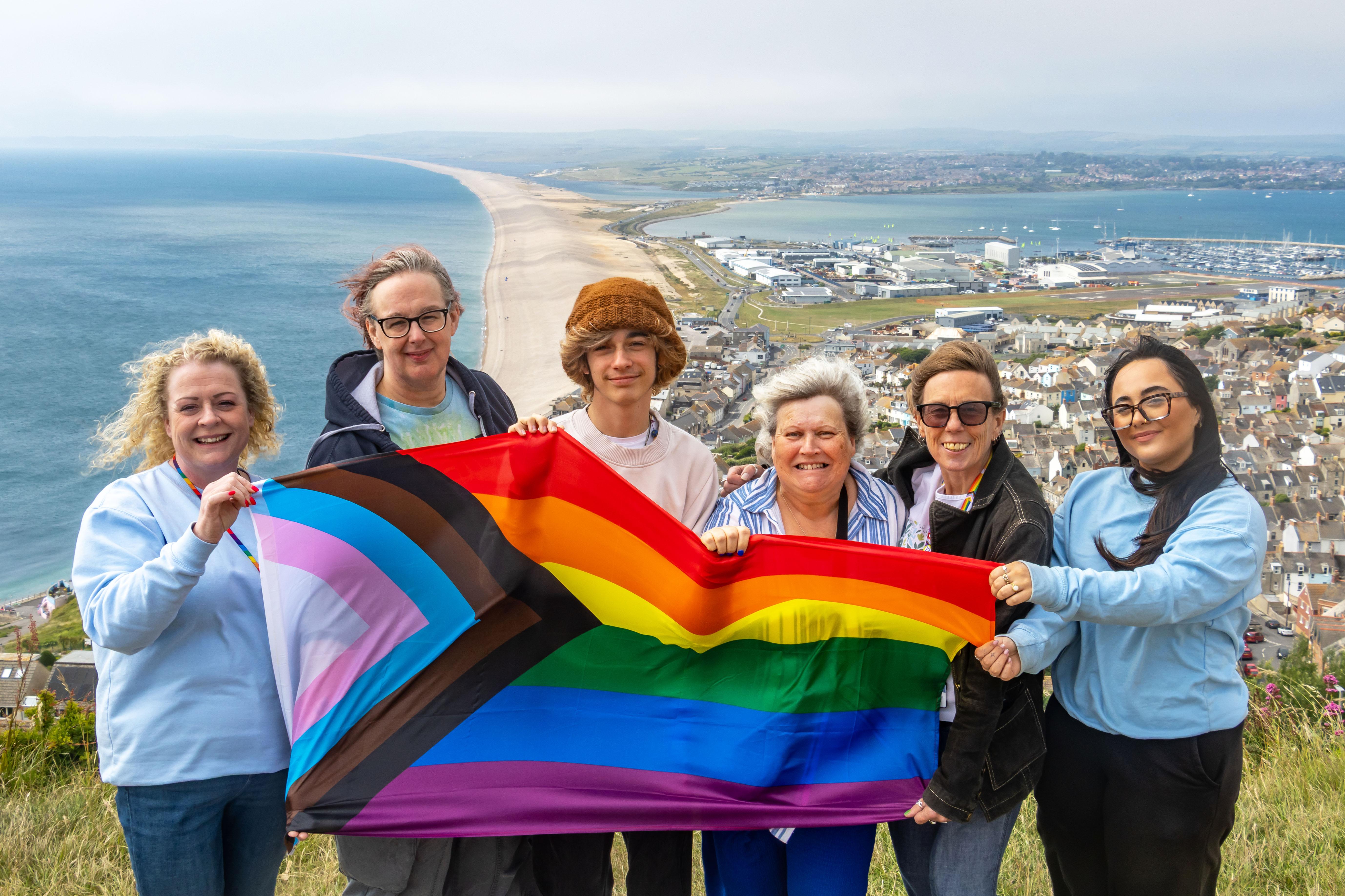 Five women and a teenage boy holding the Pride flag above Chesil beach