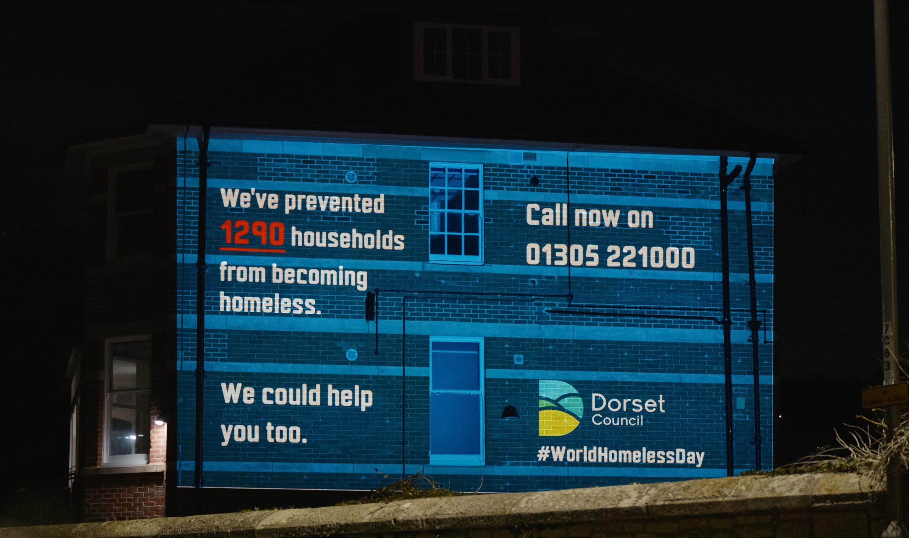The side of a red brick building has been illuminated with a blue background with white text which has the council's housing helpline number on it with their logo and hash tag world homeless day