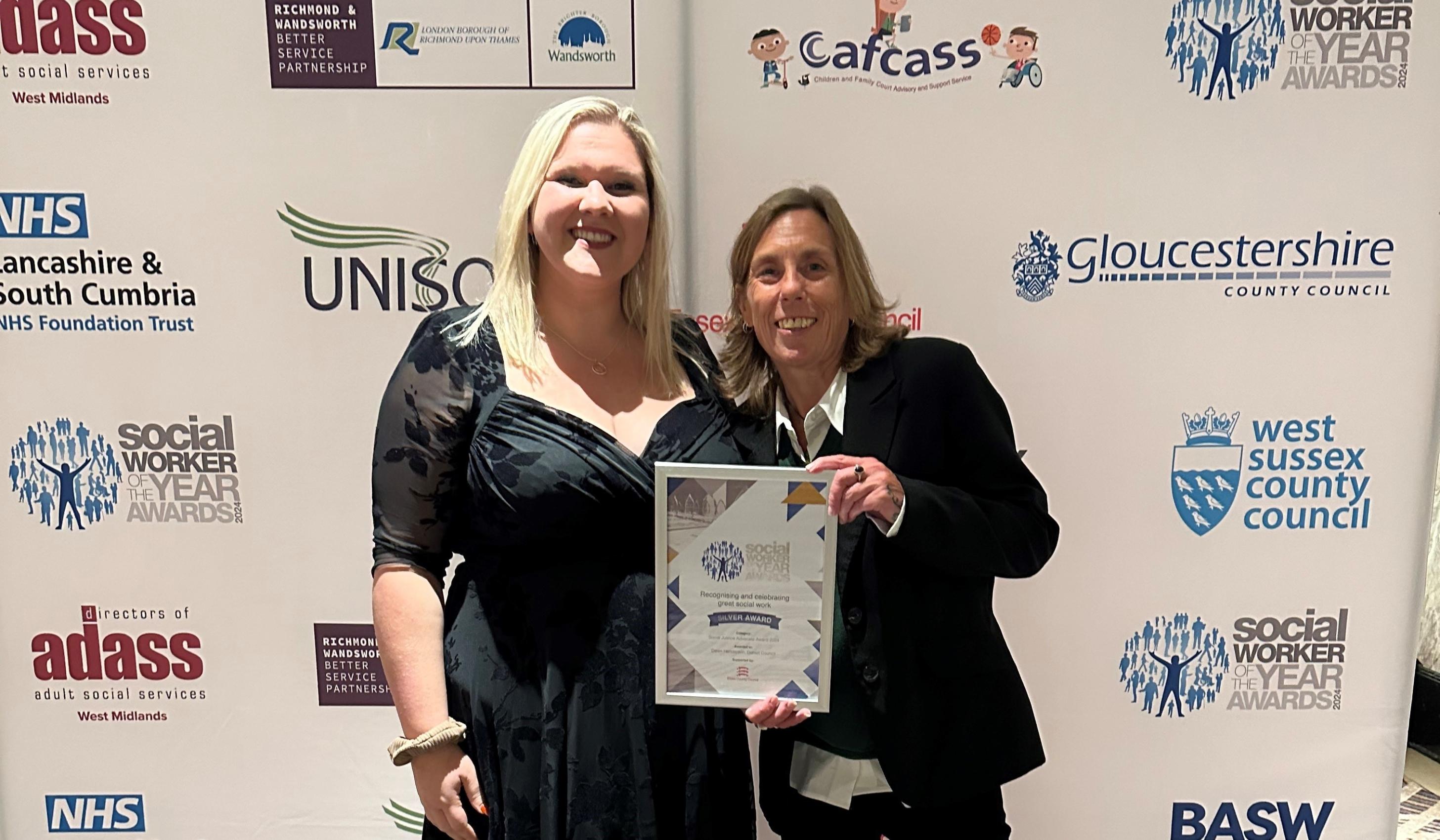 two people standing in front of a board with company logos. One of them is holding an award certificate.