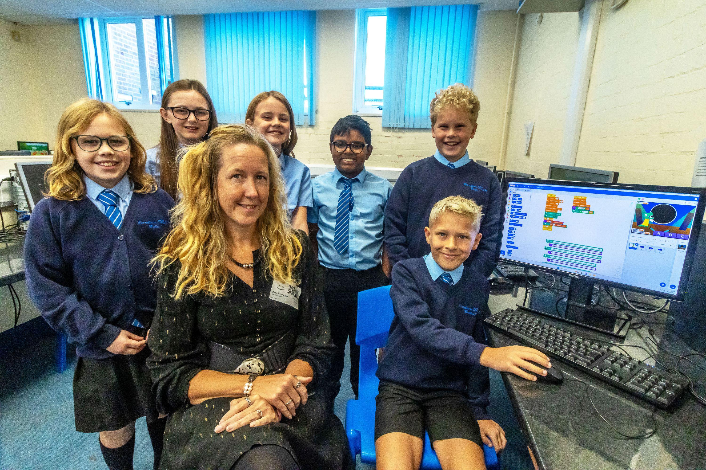 Six school children wearing navy blue uniform are sitting in a room of computers. Sitting with them is a woman with long blonde hair and wearing a black dress. She is a tech volunteer supporting the children with their Dorset Coding Week activity