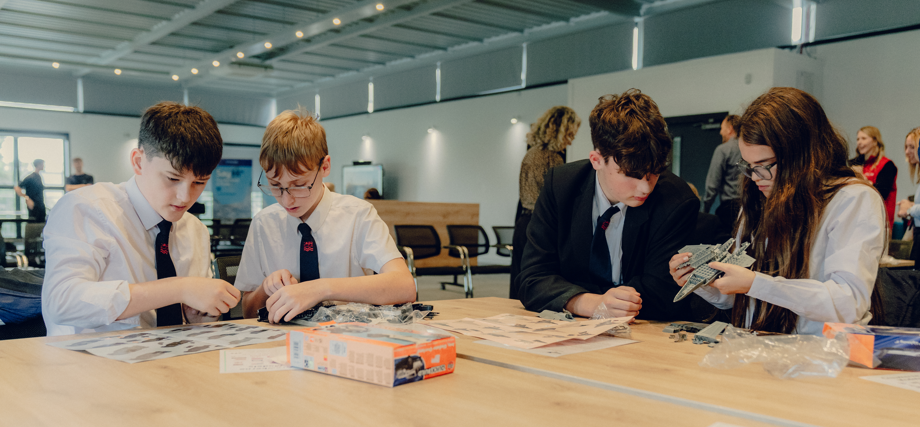 Two groups of two students who are sat next to each other at a pale coloured table are doing Air Fix models of aeroplanes in a large room.