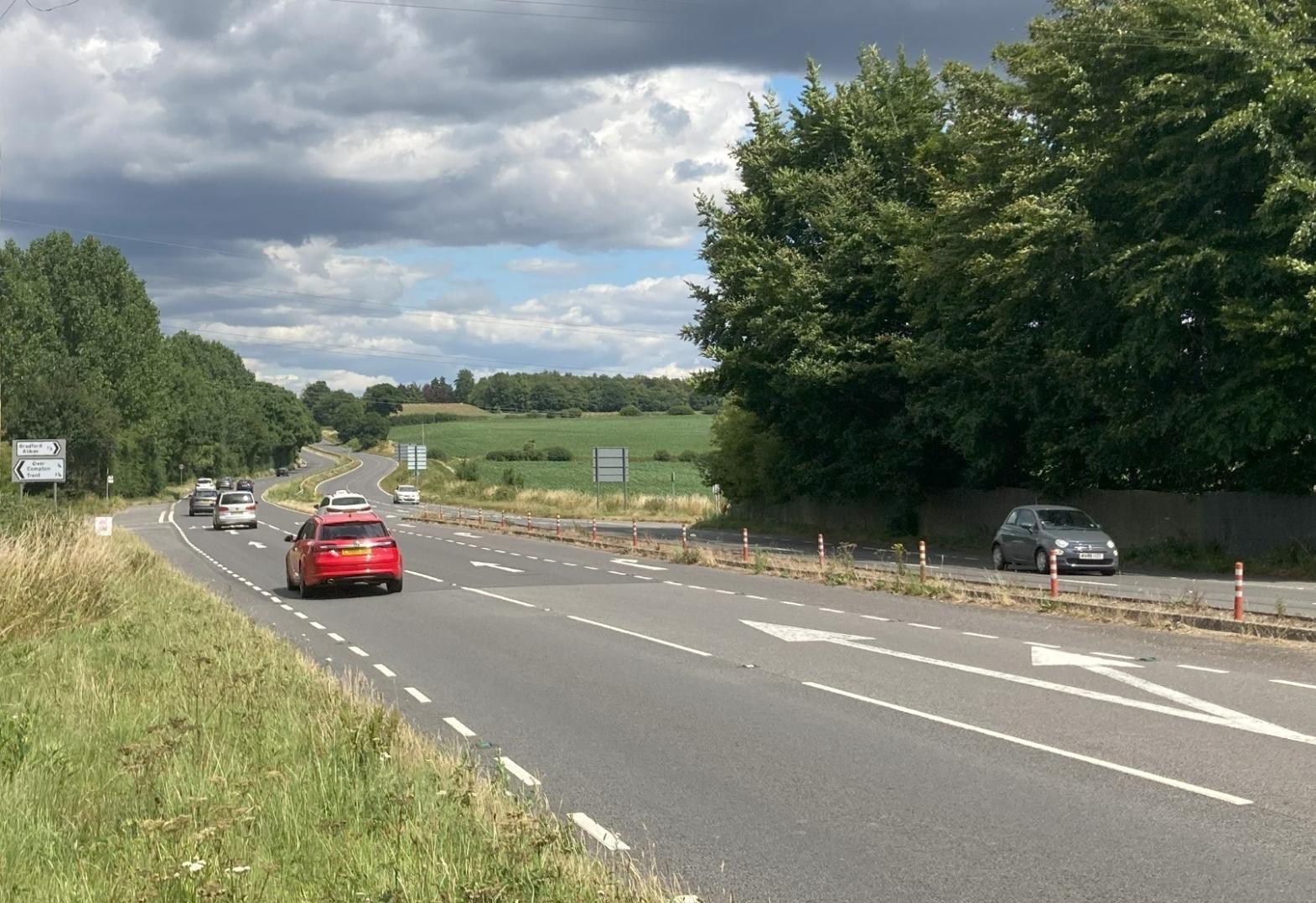 A road with broken white lines. There are arrows on two of the sections and cars, including a red car, a white taxi and a silver car heading in east. There is a grey car travelling west on the other side of the carriageway.