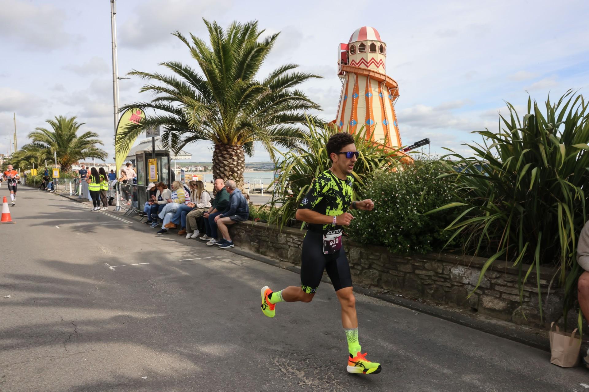 A man running beside a wall where people are seated, with a helter skelter fairground attraction and palm trees behind