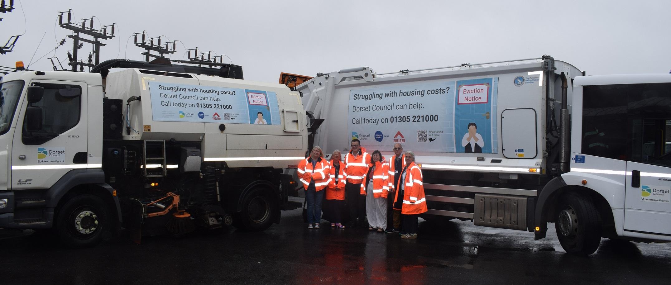 6 people are wearing orange hi-vis jackets stood in front of 2 waste collection vehicles which are parked at an obtuse angle.