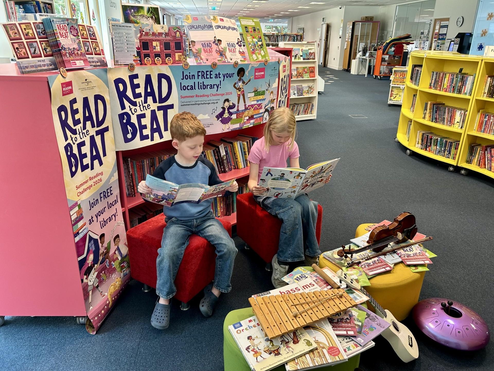 Two children reading in a colourful library surrounded by books and musical instruments with the READ to the BEAT posters behind them