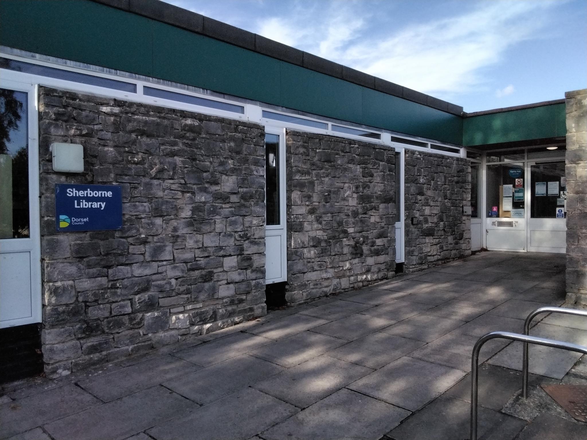 Exterior of Sherborne Library, showing a low stone‑walled building with a Dorset Council sign by the entrance and a paved walkway leading to the front doors.