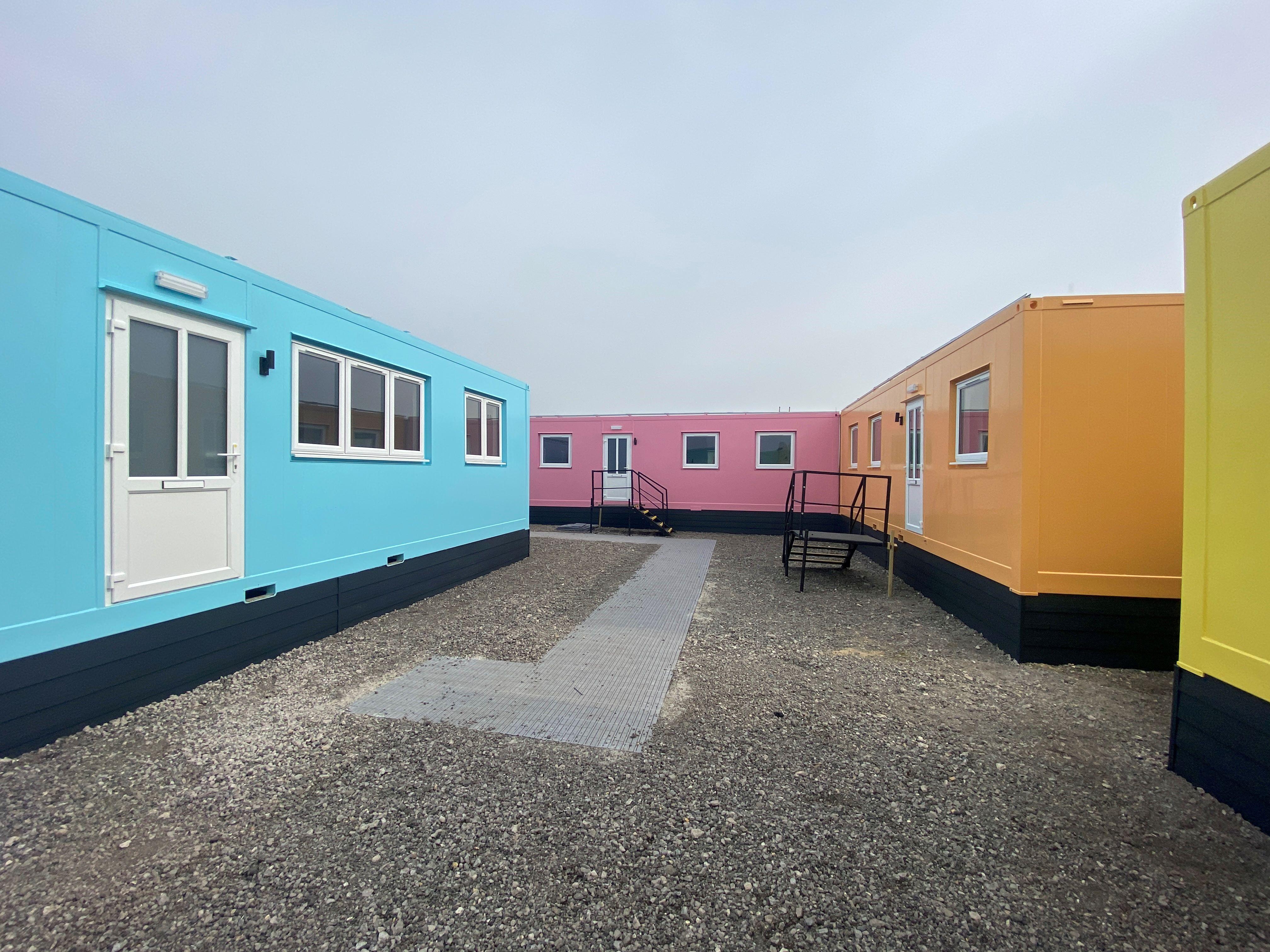 Single storey multicoloured modular units of accommodation are in a U shape on a gravel surface. The units are sky blue, pink, orange and yellow.