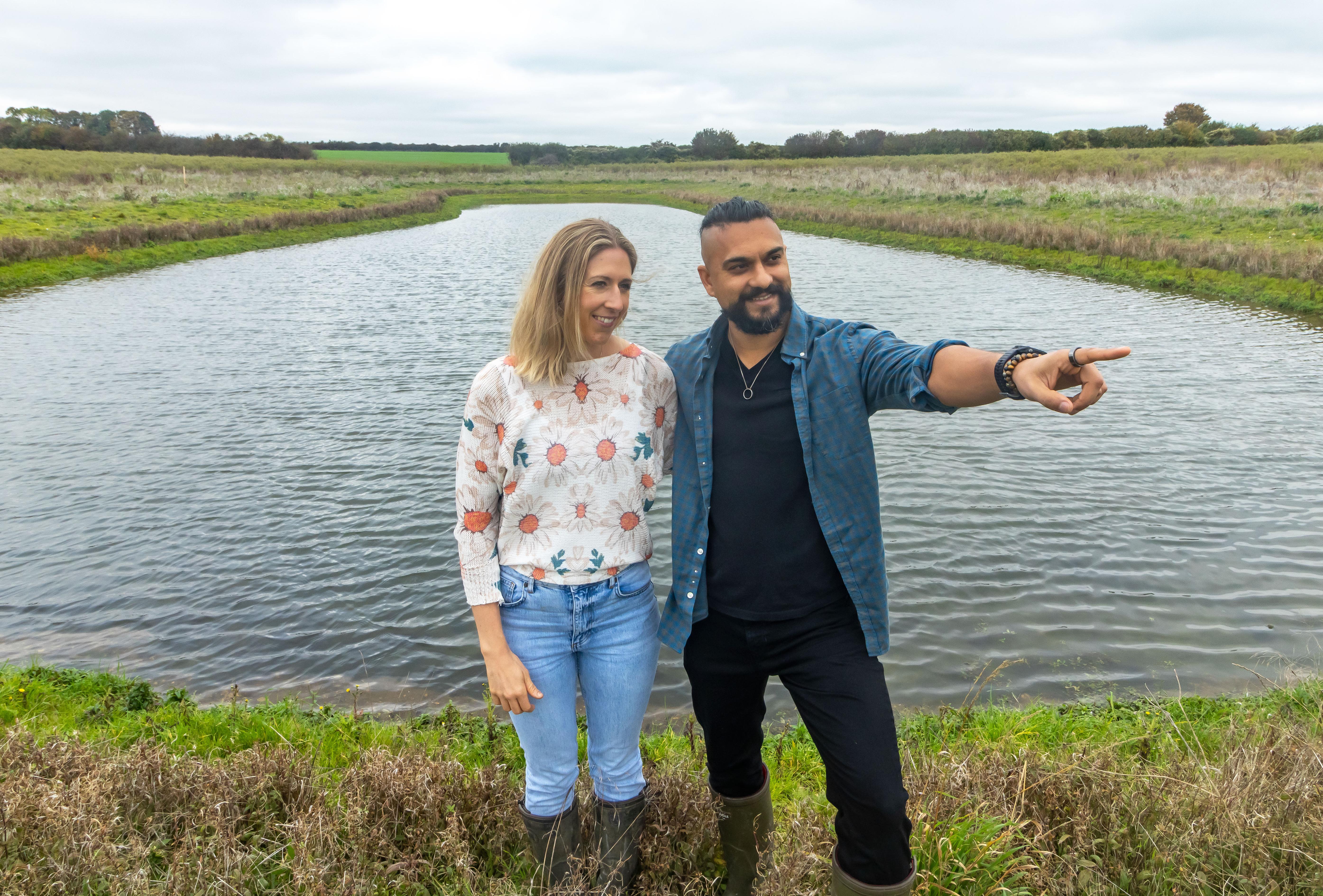 A woman with blonde hair and wearing jeans, wellies and a jumper is standing next to a man with black hair and a black beard, he is wearing a blue shirt, black trousers and wellies. They are both standing in front of a large pond surrounded by countryside