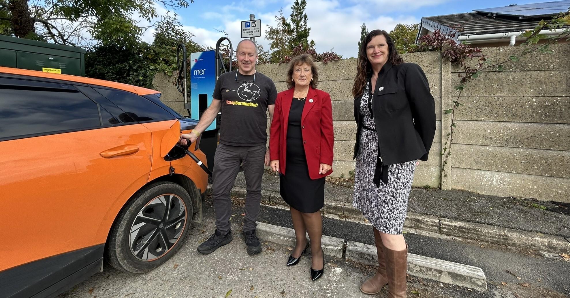 three individual pose for picture next to electric vehicle and charge point, one of the individuals is charging a car.