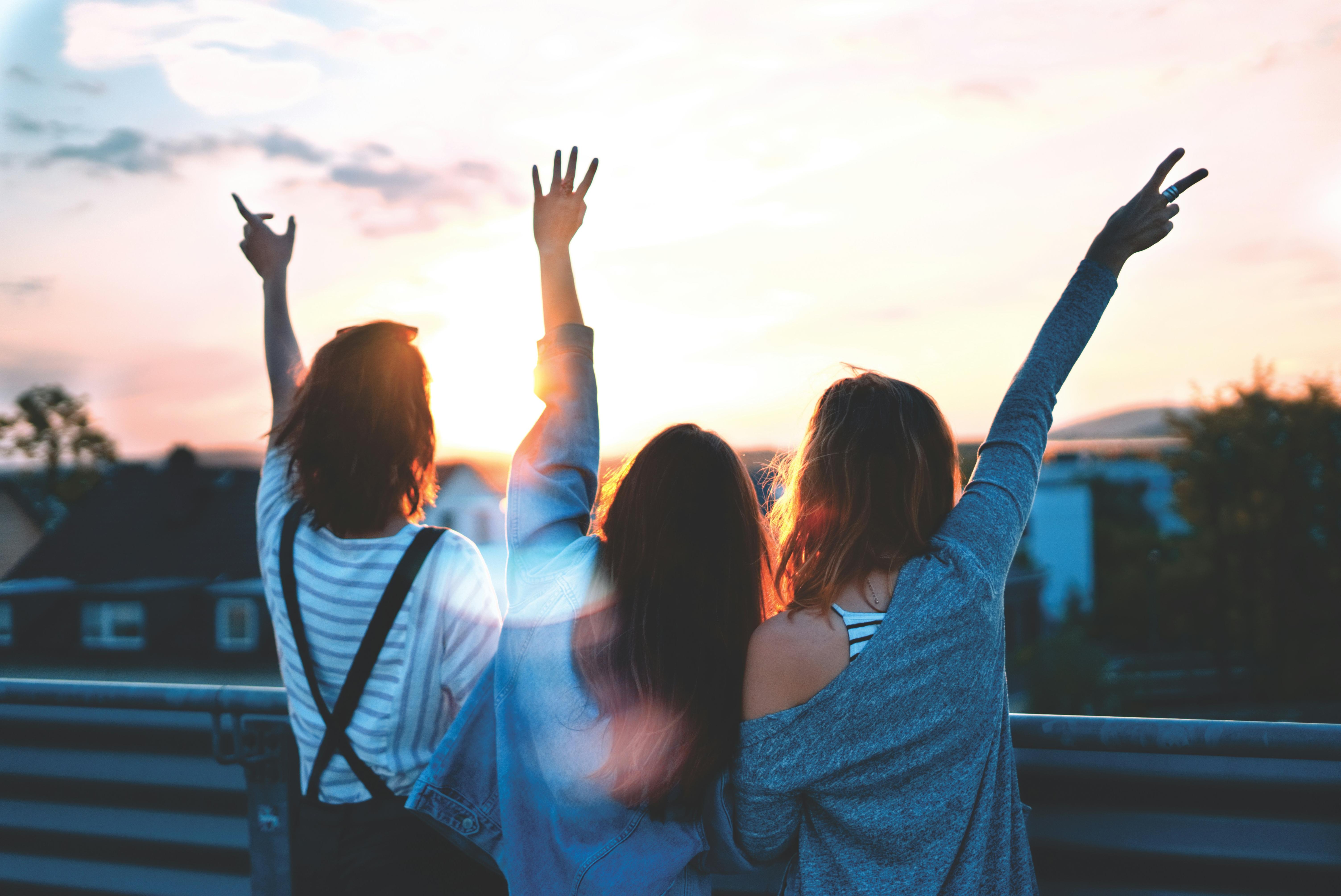 three young women with their backs to the camera and hands in the air looking at a sunrise