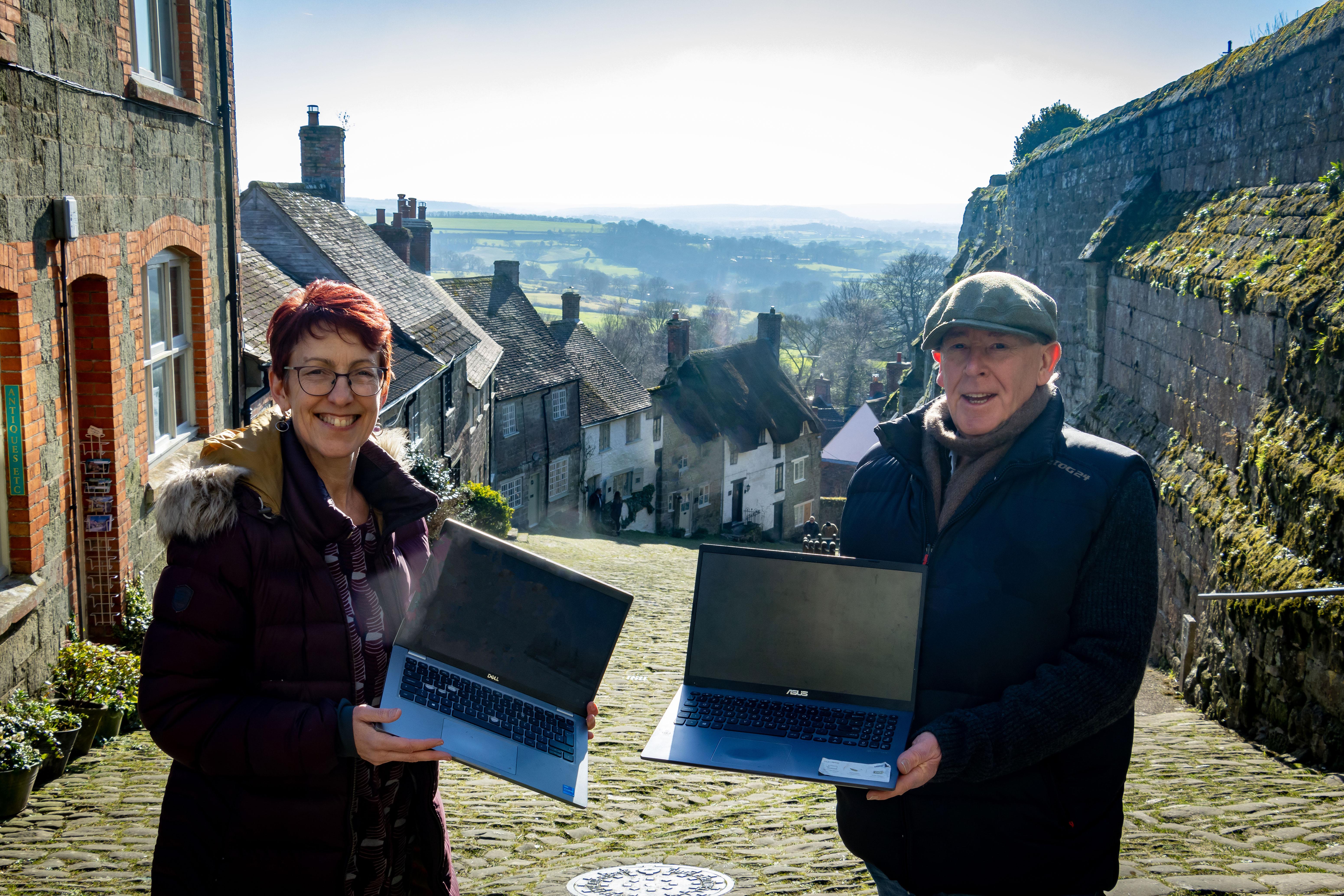 A woman with short red hair and glasses is holding a laptop next to a man wearing a flat cap, who is also holding a laptop. They are standing on a cobbled path with thatched cottages in the background. This is Gold Hill in Shaftesbury.