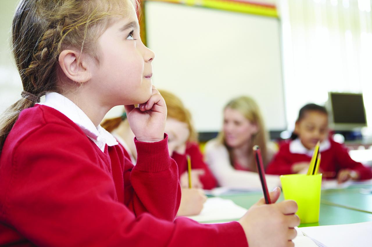 Children in red jumpers sat around a table with pencils in their hands. It looks like a school classroom.