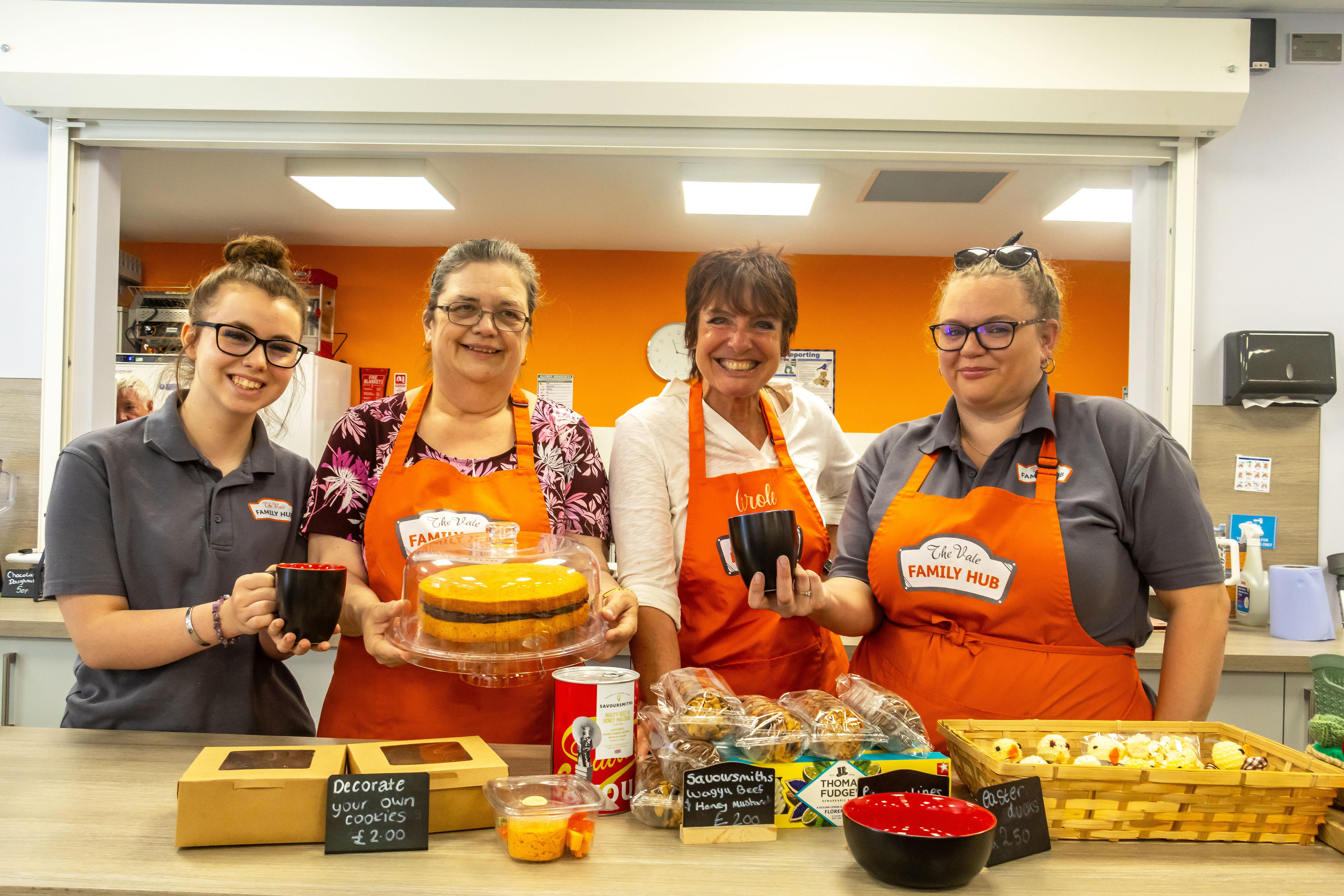 Four women wearing orange aprons are in a cafe holding mugs of coffee and cakes