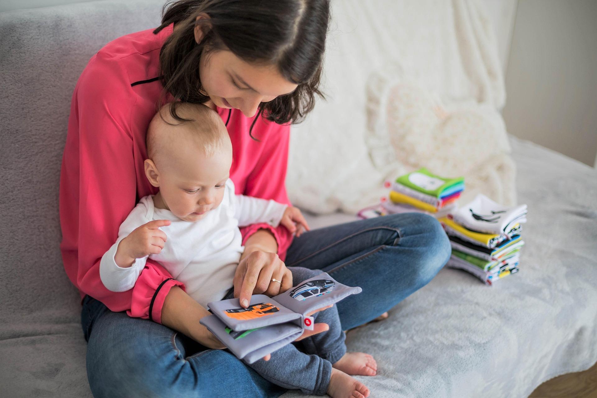 A white young woman has a white baby sat on her lap on a light coloured sofa. She is cuddling the baby. They are reading a soft book together.