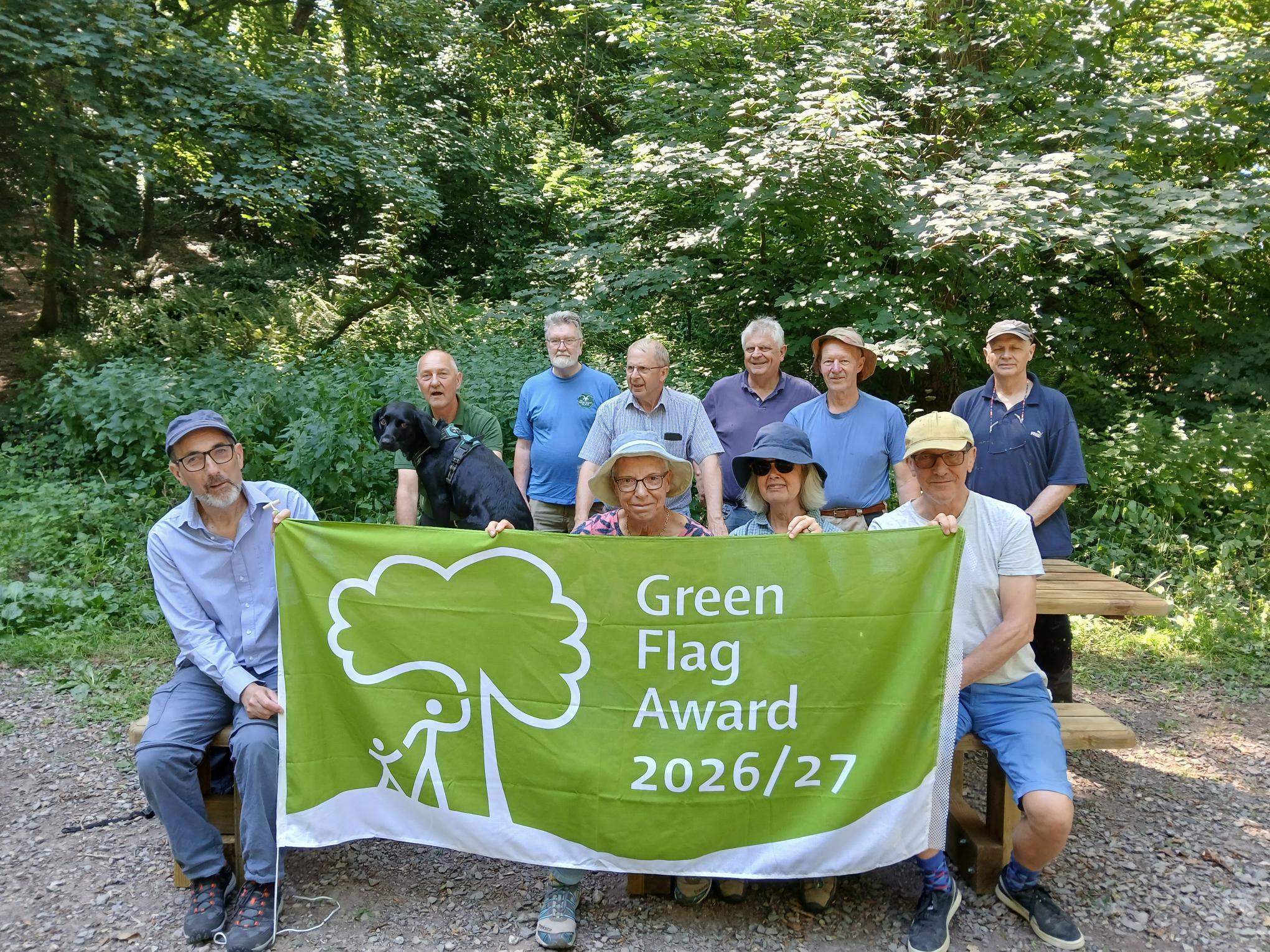A group of 10 people and one black dog pose in a woodland clearing, holding a large green flag that reads “Green Flag Award 2026/27”. The group stands and sits around a picnic bench surrounded by dense green trees and vegetation. Sunlight filters through.