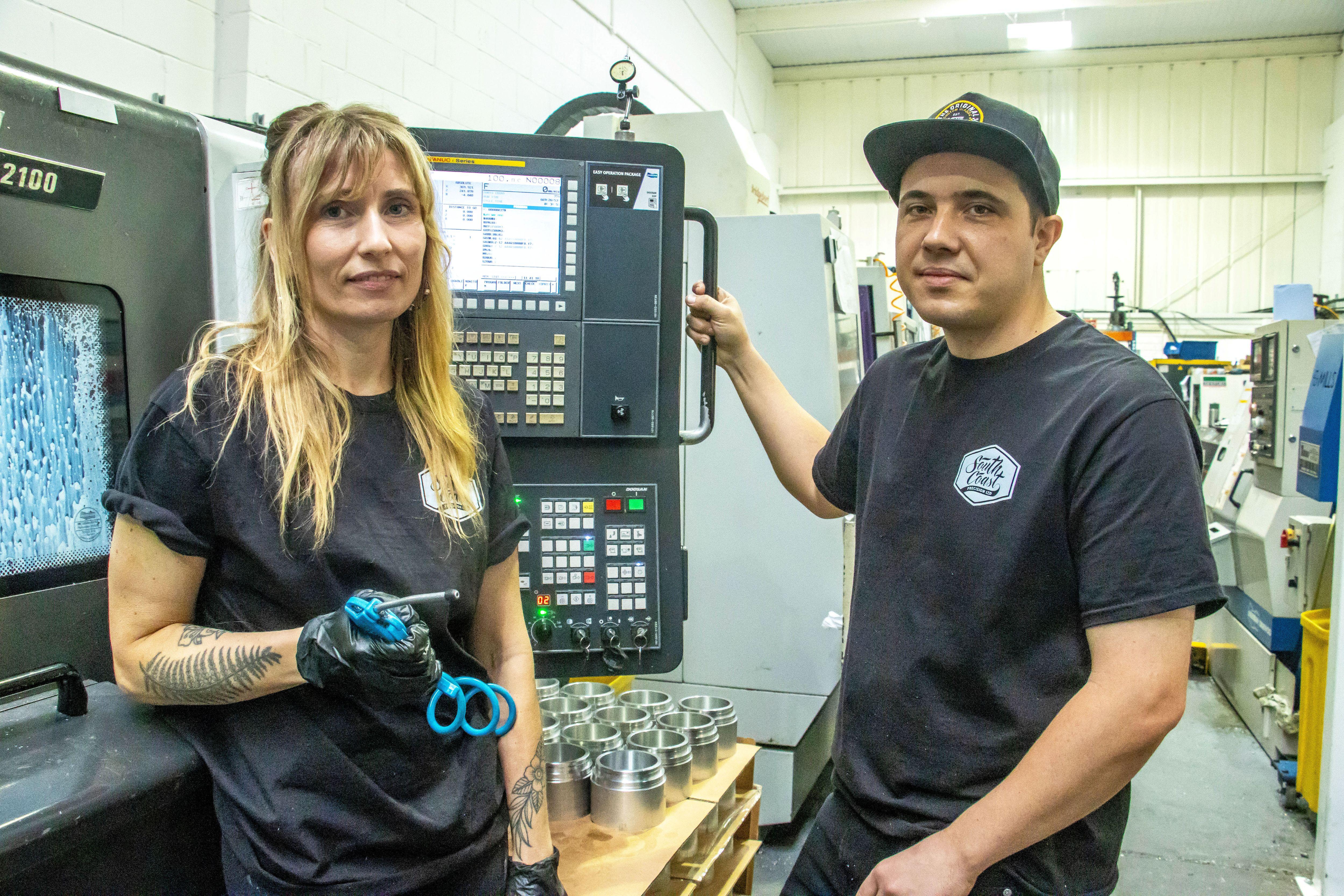 A woman with long blonde hair and wearing a black t-shirt is standing next to a man wearing a baseball cap and black t-shirt. They are both standing in front of a lathe machine.