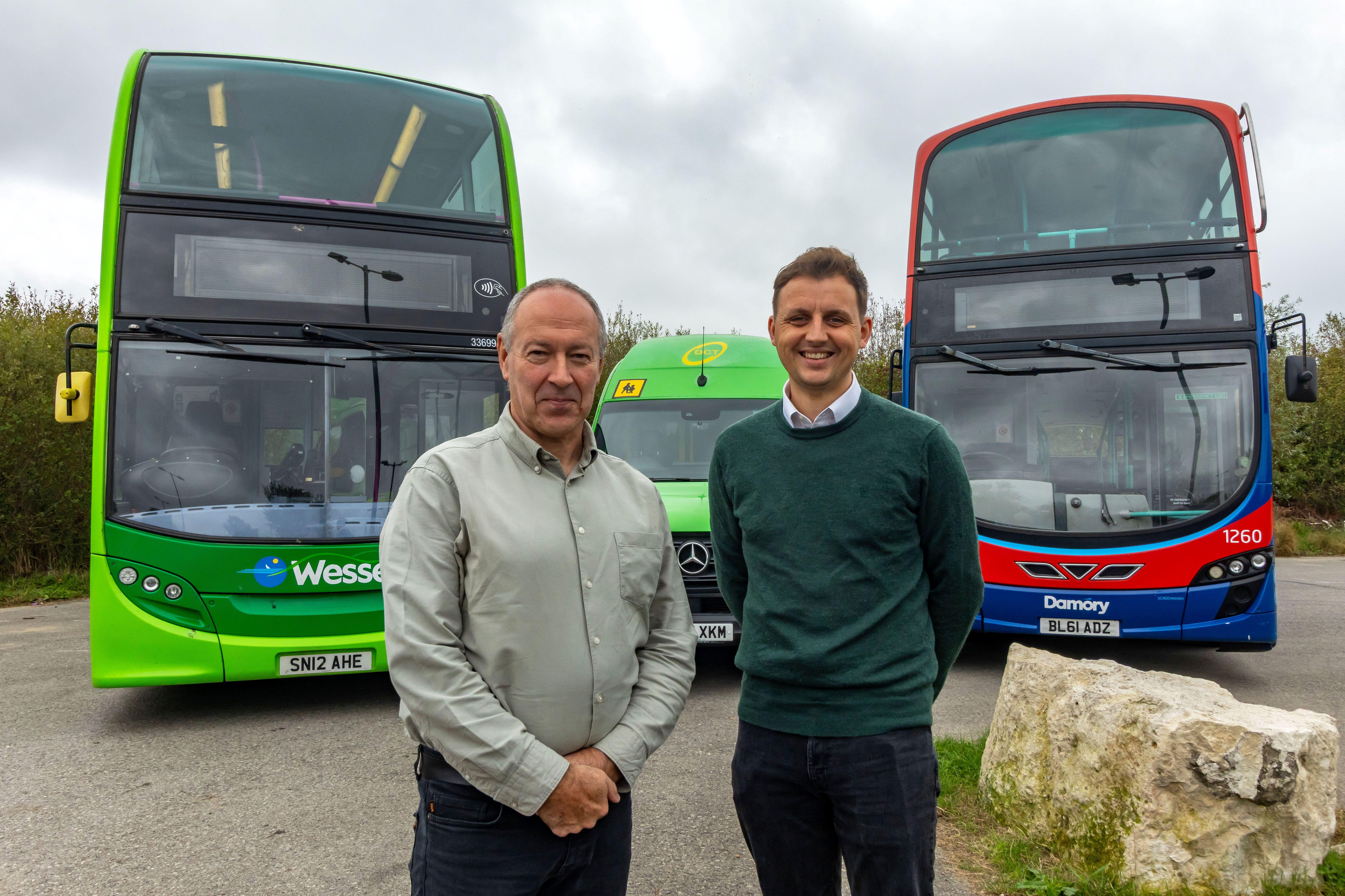 Two men standing in front of 2 double decker buses and a mini bus