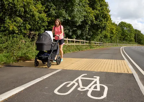 Person pushing a pram along a shared-use path marked for cycling, beside a road and bordered by trees.