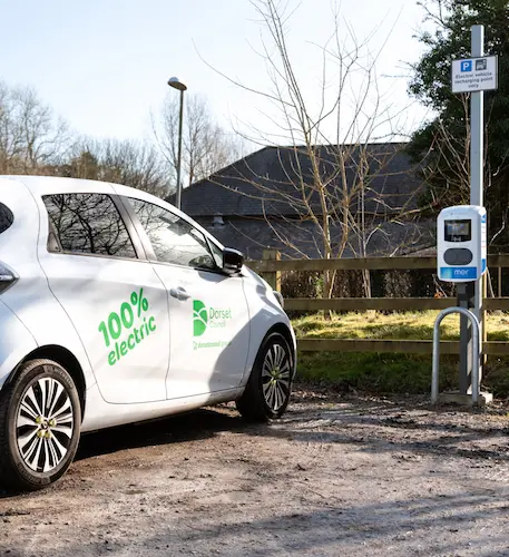 Electric vehicle parked beside a public charging point, with signage for electric vehicle charging and trees and buildings in the background.