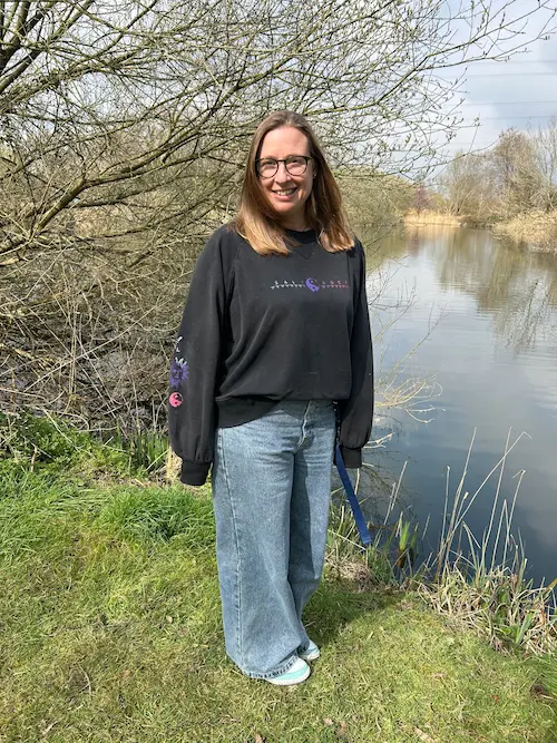 Picture of Anna standing on a grassy riverbank beside a calm waterway, with trees and reeds in the background.