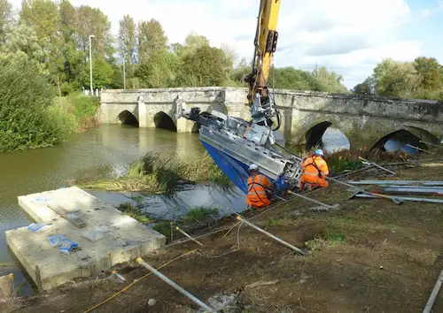 Construction activity along the riverbank at Sturminster Newton, with visible reinforcement structures and machinery. The image highlights engineering works to stabilise the bank and safeguard the A357 against erosion and flooding. 