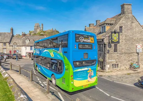 A bus travelling along a country road toward the village of Corfe Castle, with the historic castle ruins visible in the background. The image highlights rural connectivity and the role of public transport in linking communities and heritage destinations.