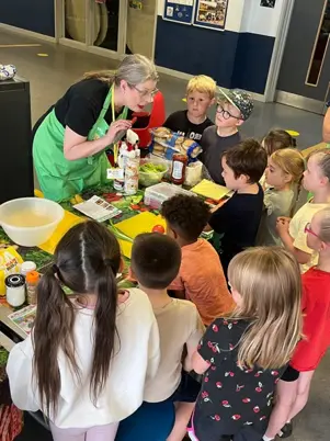 Children gathered around a table during a food activity, with ingredients, recipe materials and healthy foods displayed while an instructor leads the session.