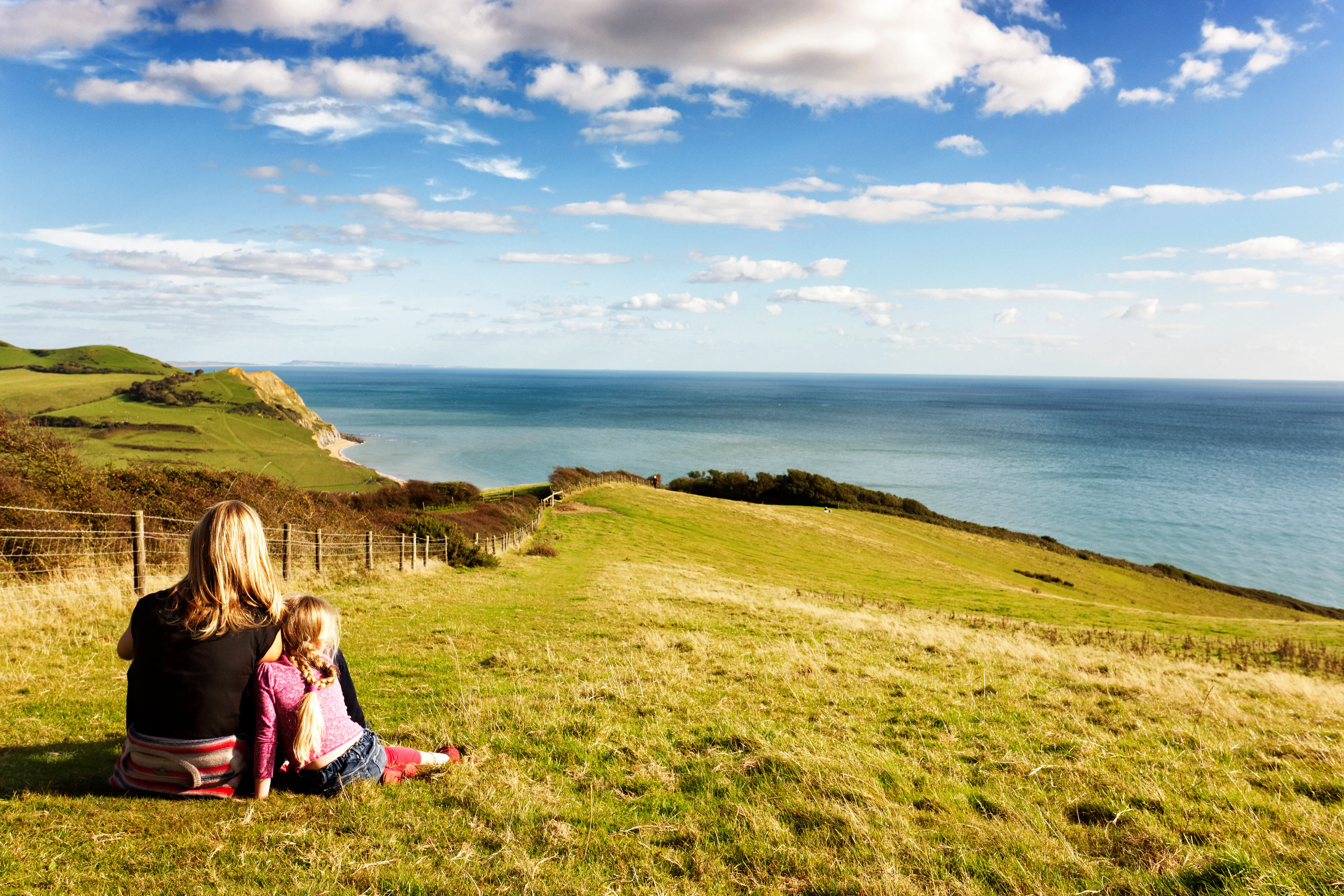 An adult and child sit on a Dorset cliff top overlooking the sea on a sunny day