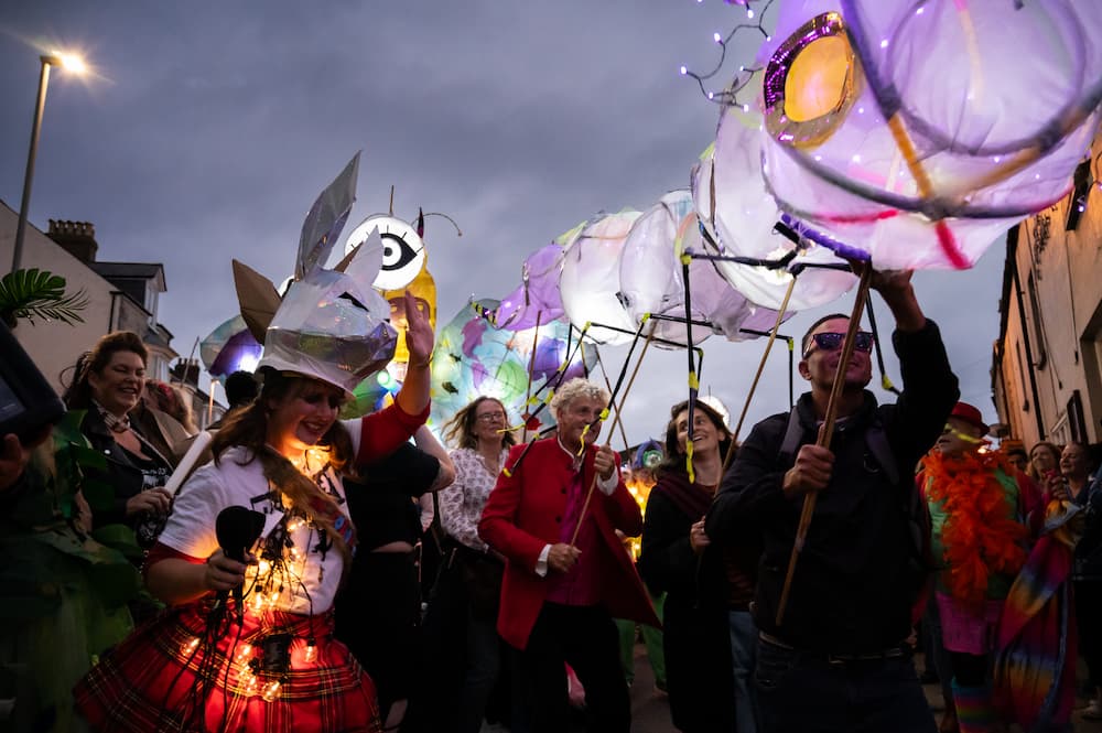 A crowd of people holding brightly coloured paper sculptures walking at sunset