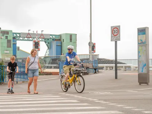 Cyclist, pedestrian and child on a scooter using a crossing near Poole Quay, with the lifting bridge and harbour infrastructure in the background.