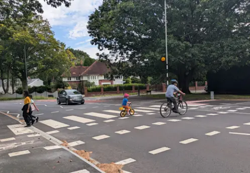 Adult and child cyclists cross a signal-controlled cycle crossing on a residential street, with a pedestrian and a car nearby.