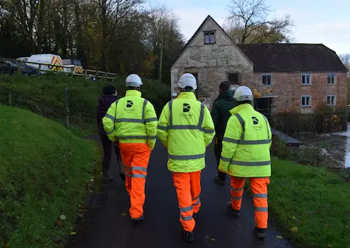 Engineering team in high-visibility clothing walking along a path during a site visit, with a stone building, trees and a river in the background.