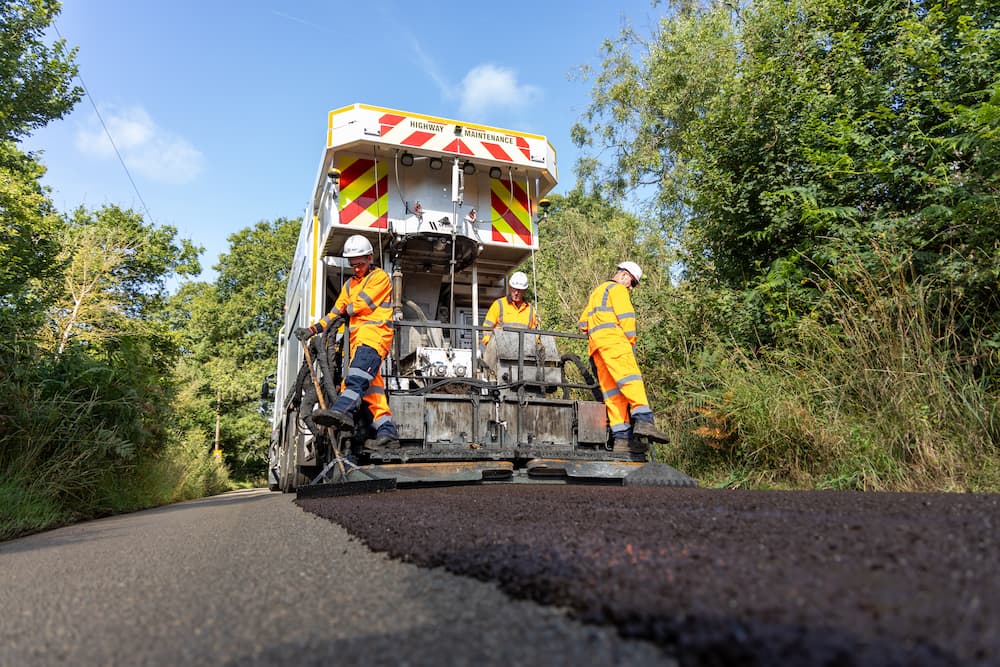 A lorry resurfacing the road with men standing on the side.