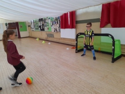 A young girl kicking a small football into a goal, with a young boy standing in front of it. They are in a sports hall.