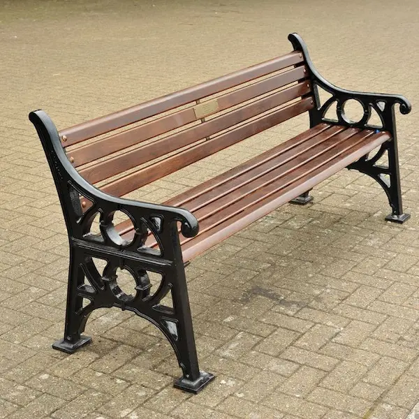 Park bench with horizontal brown slats and ornate black metal sides, positioned on paving in an outdoor public space.