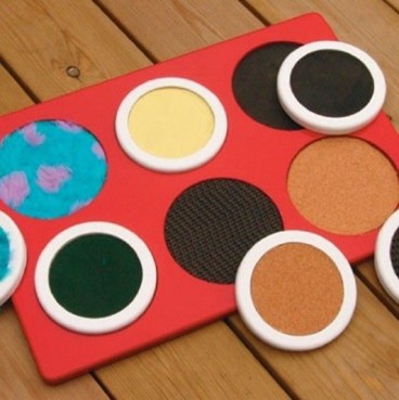 A red tactile board with pairs of textured circular pads, including soft, smooth, rough, cork and patterned surfaces.
