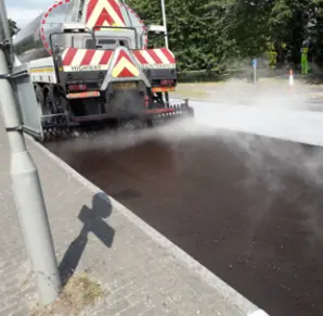 Road resurfacing work taking place on a street, with a highway maintenance vehicle spraying a fresh layer over the road surface beside a pavement.