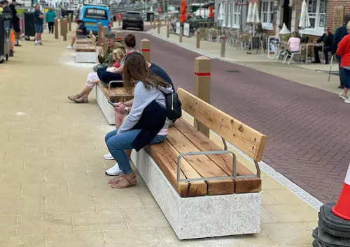 People sitting on wooden benches along a waterfront street, with a wide pedestrian area, outdoor cafés and a roadway running alongside.